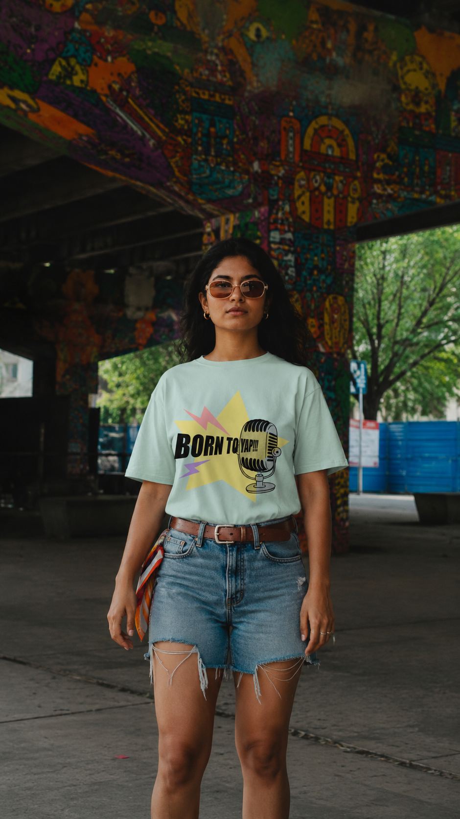 A woman wearing a light mint green graphic t-shirt with a 'BORN TO BE' microphone design, paired with denim shorts, standing under a colorful graffiti-covered bridge.