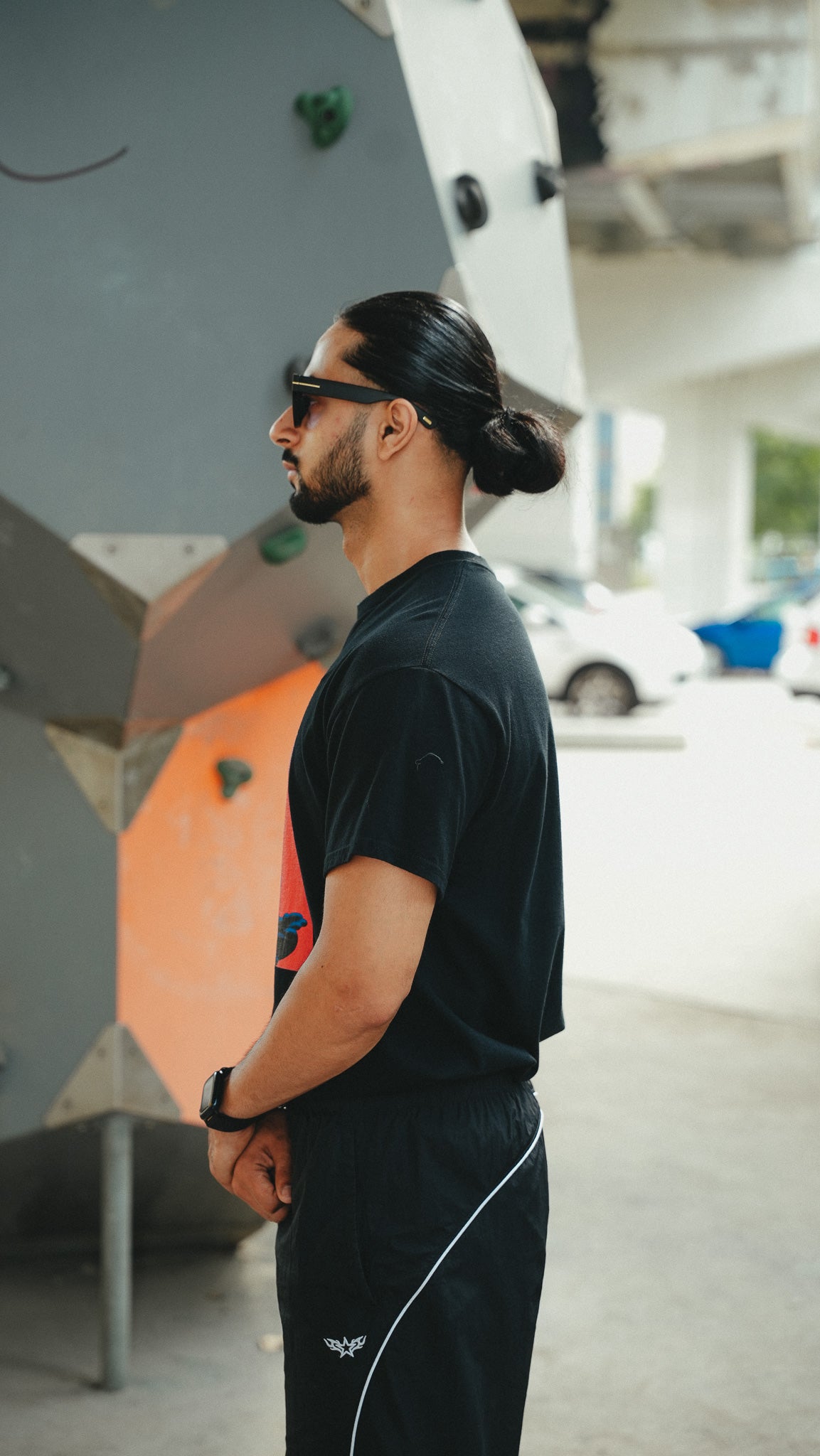 A man in a black t-shirt and black track pants with a white stripe stands in profile outdoors next to a climbing wall.