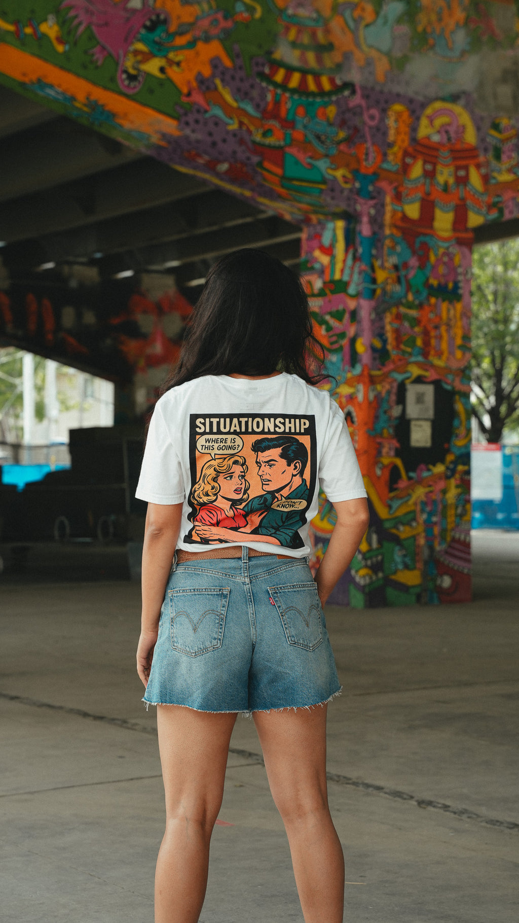 A woman stands with her back to the camera wearing a white graphic t-shirt with a vintage-style comic book illustration and the word 'SITUATIONSHIP' printed on the back, paired with denim shorts in an outdoor underpass with colorful graffiti.