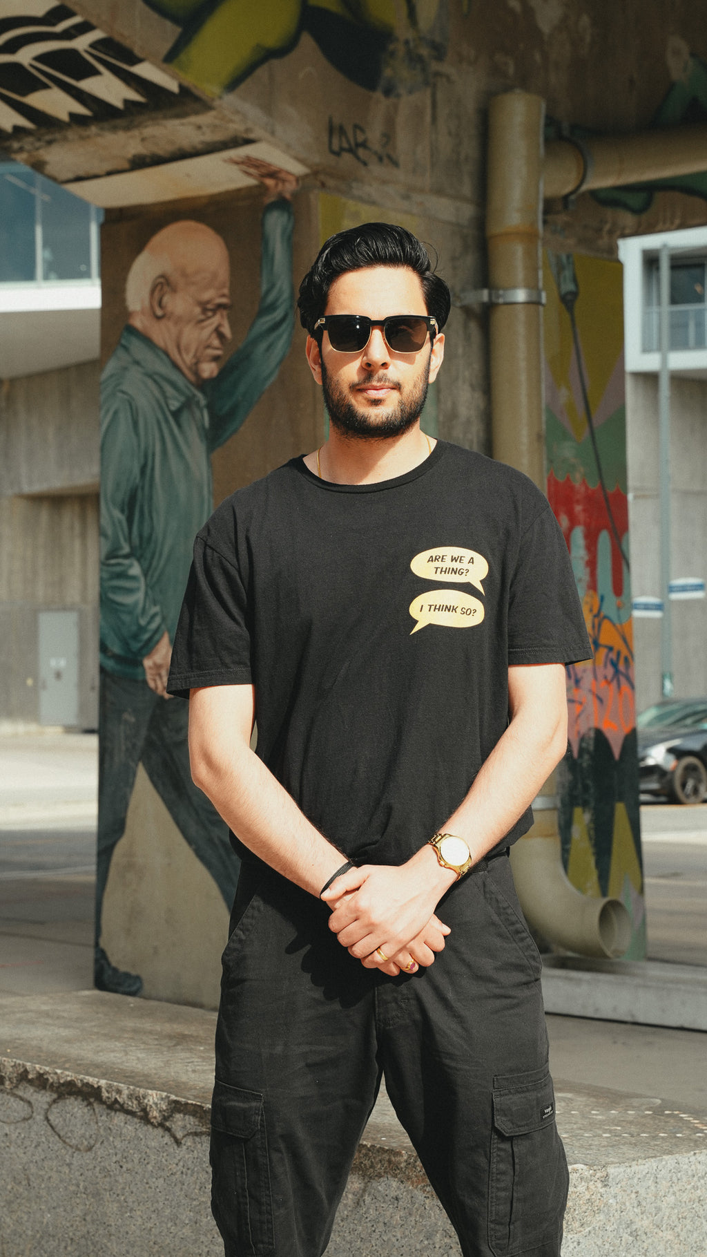 A man wearing a black graphic t-shirt with two speech bubbles and black cargo pants, standing outdoors in front of a concrete wall with a mural.