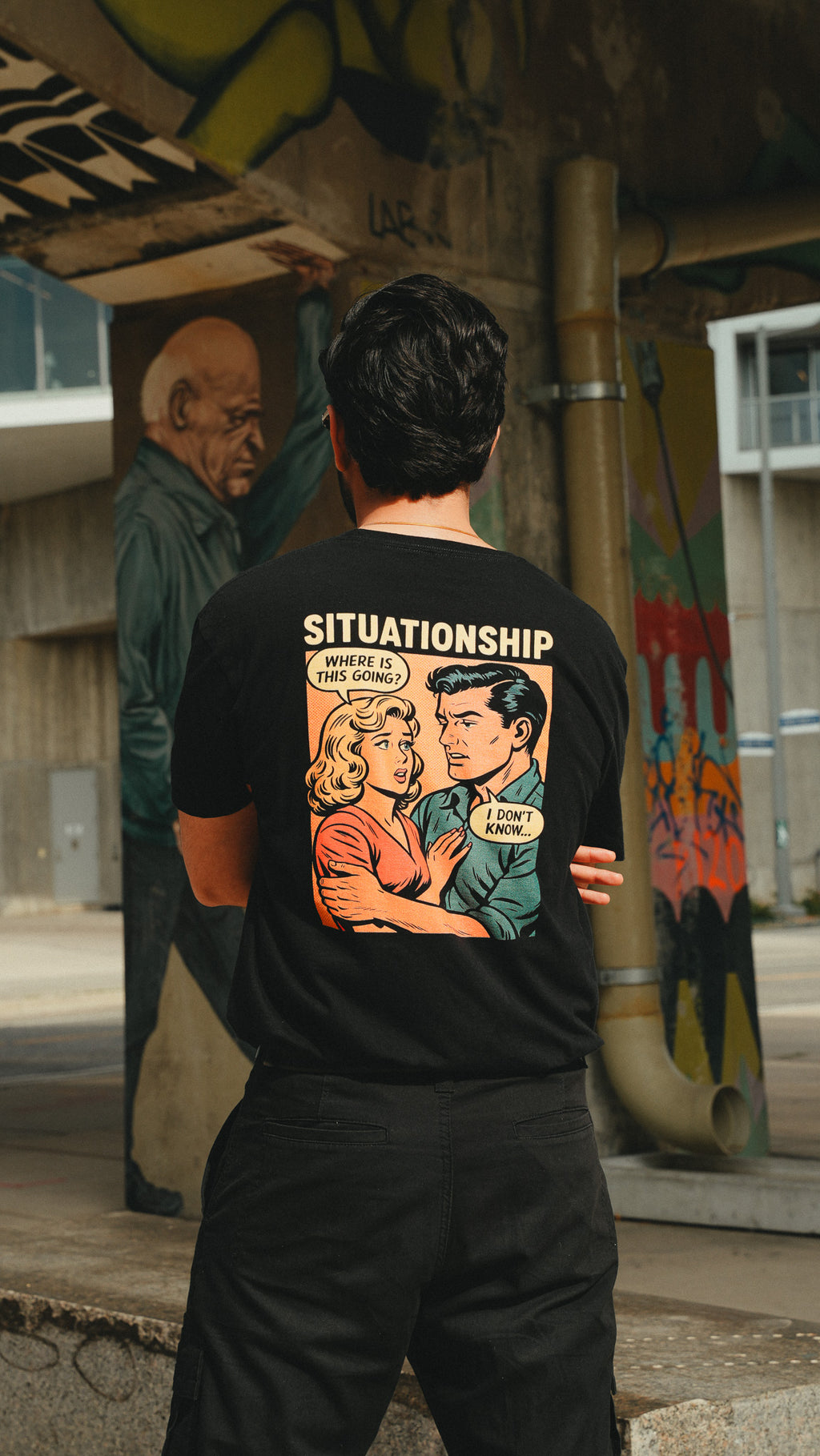 A man viewed from behind wearing a black t-shirt with a large retro comic-style graphic on the back. The graphic features the text 'SITUATIONSHIP' and an illustration of a couple with speech bubbles.