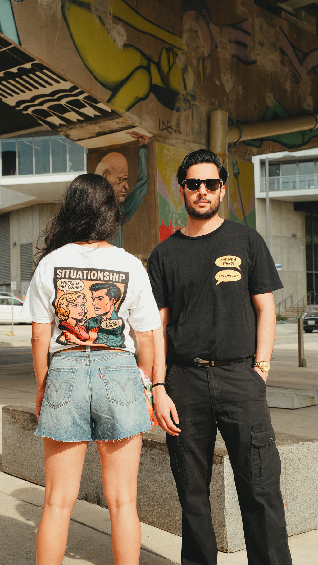A man and a woman standing outdoors in front of a graffiti-covered concrete structure. The woman is shown from the back wearing a white graphic T-shirt with a cartoon-style illustration and the word 'SITUATIONSHIP' on the back, paired with denim shorts.