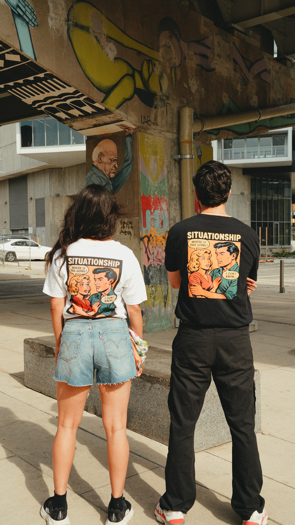 Two people standing under a graffiti-covered bridge, viewed from behind. The woman wears a white graphic T-shirt and denim shorts, and the man wears a black graphic T-shirt and black pants.
