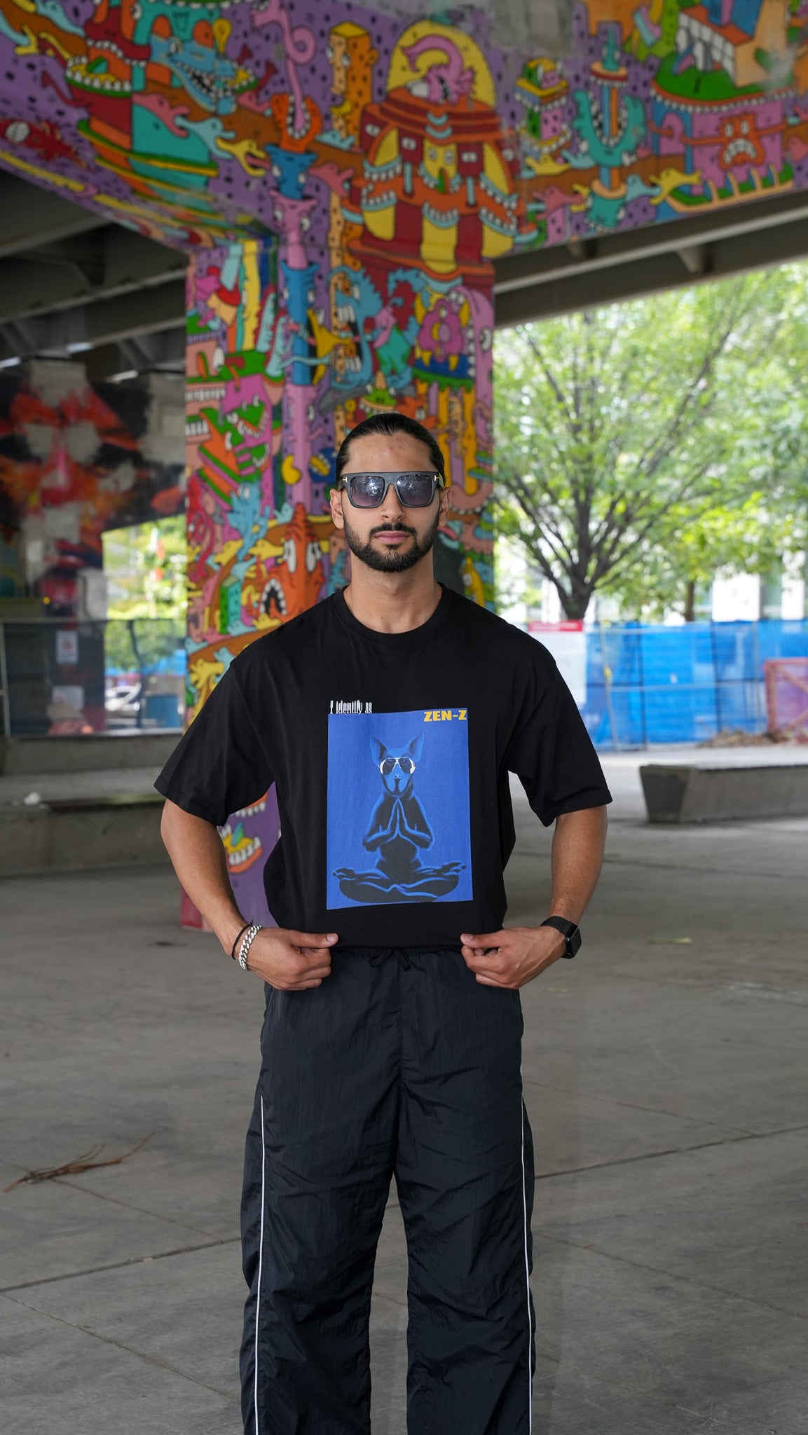 A man wearing a black graphic t-shirt with a blue and black print, paired with black pants, standing in an outdoor urban setting under a colorful graffiti-covered bridge.