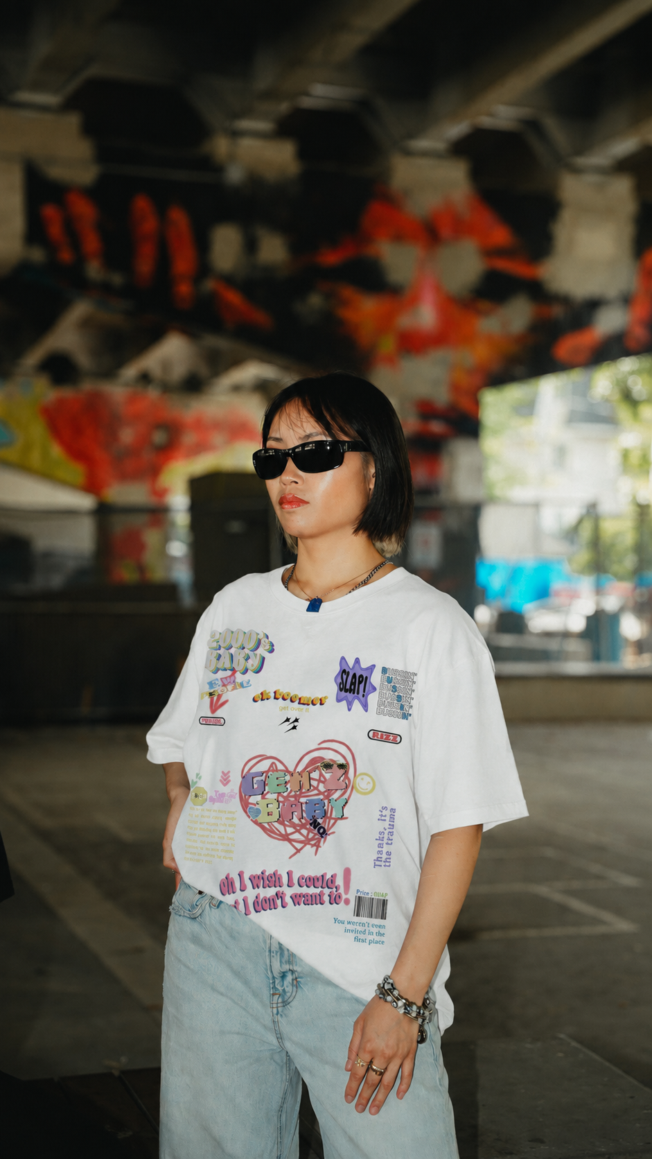 A person wearing a white graphic t-shirt with colorful cartoon and text prints, paired with light blue jeans and black sunglasses, standing in an urban outdoor setting with graffiti walls in the background.
