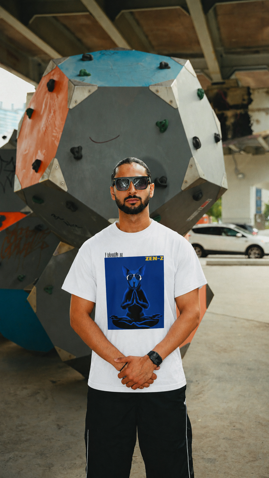 A man wearing a white graphic t-shirt with a blue meditating figure print, black sweatpants, and sunglasses, standing in front of a large geometric climbing wall structure.