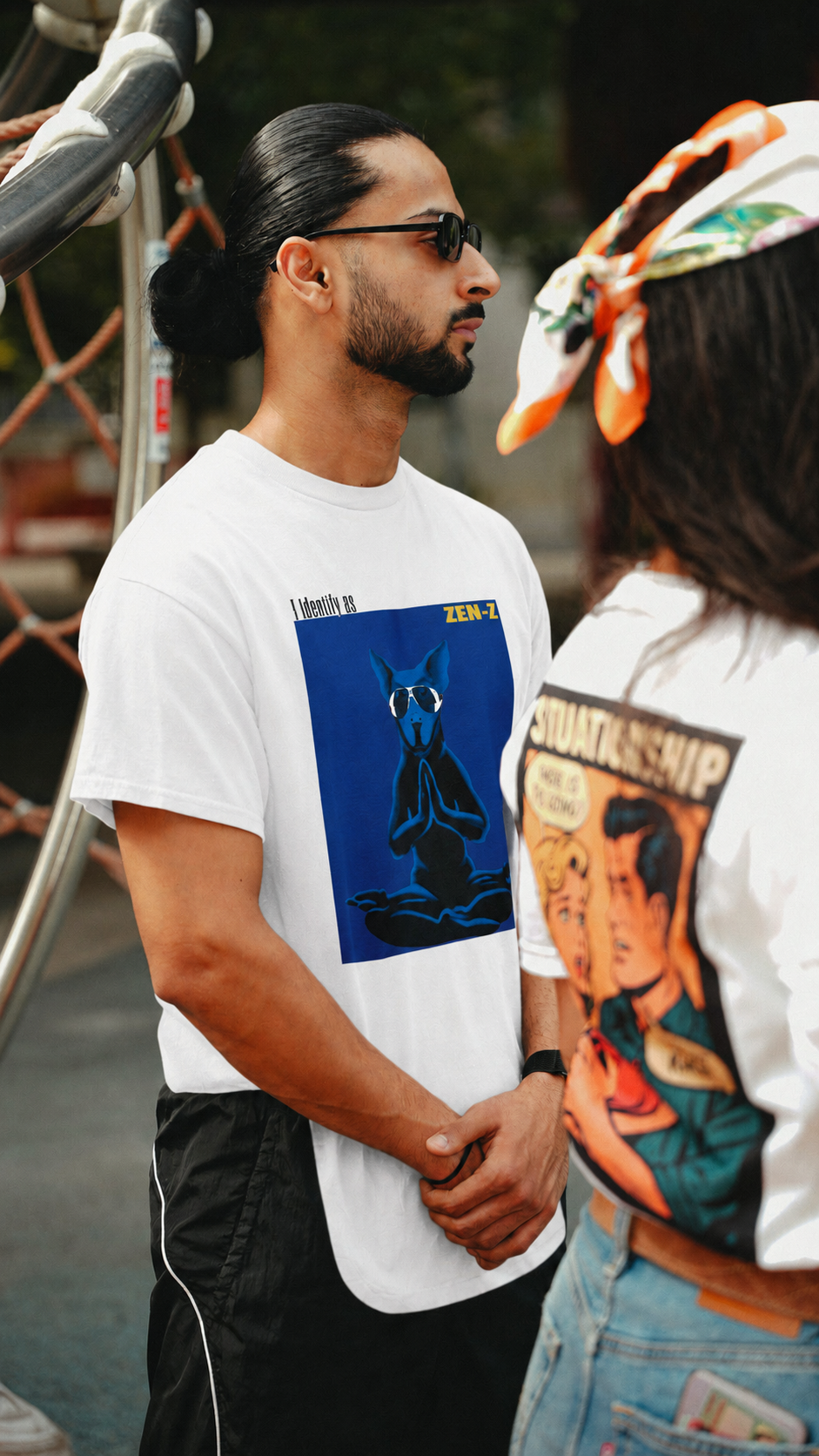 A man wearing a white graphic t-shirt with a blue cat illustration stands in an outdoor setting next to another person wearing a white t-shirt with a colorful comic-style graphic.