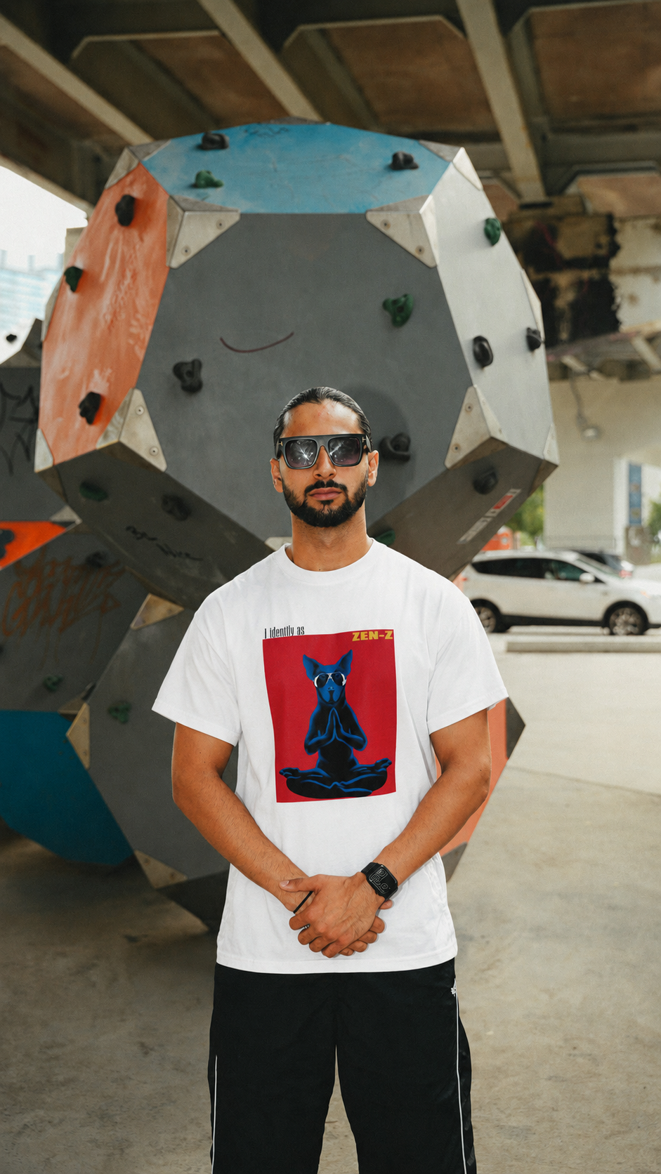 A man wearing a white graphic t-shirt with a red and blue dog print, black pants, and sunglasses, standing in front of a large geometric climbing structure.